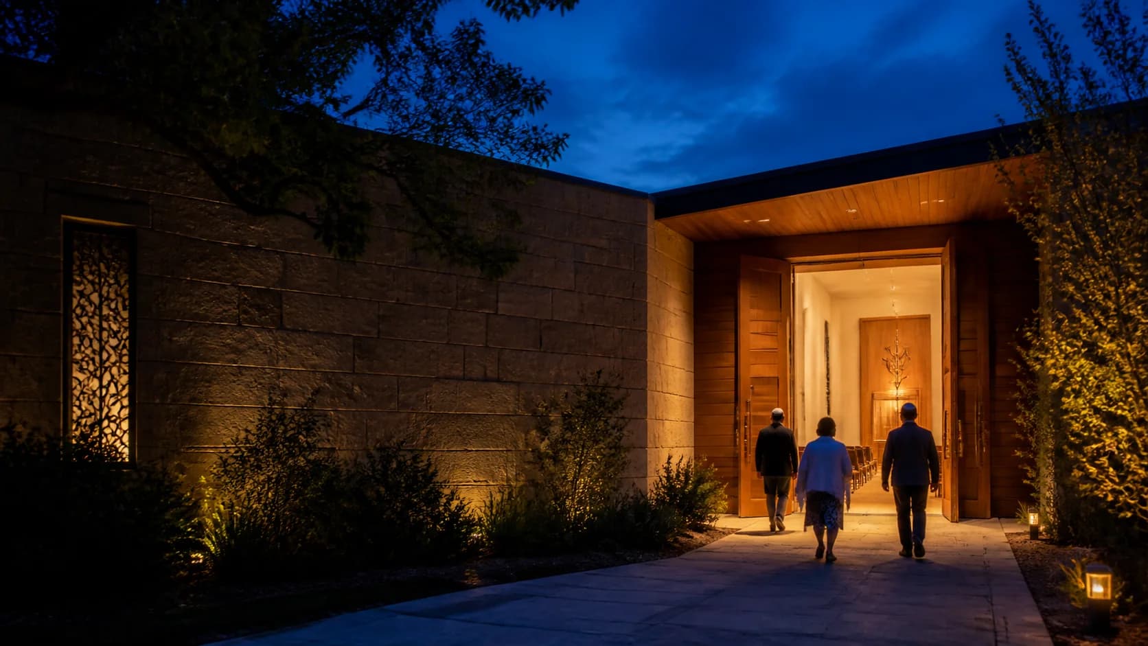 Three people walking up a lit path into a synagogue at dusk, its wooden doors open.